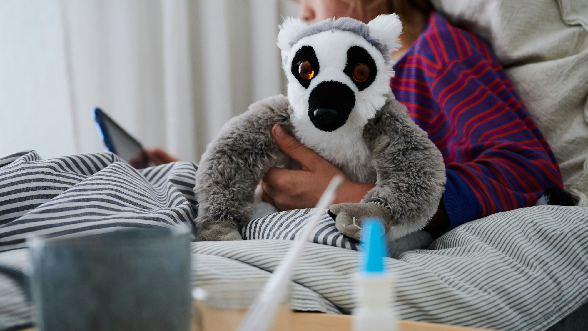 Eine Person liegt im Bett und guckt auf ihr Tablet, in der Hand ein Faultier-Kuscheltier. Auf dem Nachtisch stehen ein Fieberthermometer und ein Erkältungsspray.