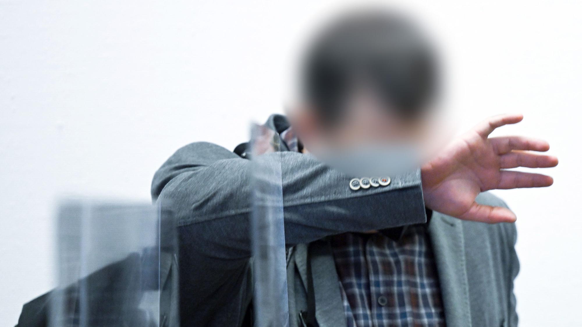 Der Angeklagte steht vor dem Beginn des Prozesses im Gerichtssaal im Landgericht und bedeckt sein Gesicht.