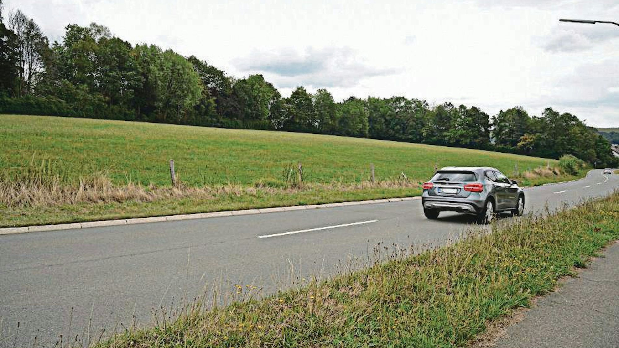 Ein Feld in Kürten. Im Hintergrund Bäume. Im Vordergrund eine Straße mit einem silbernen Auto.