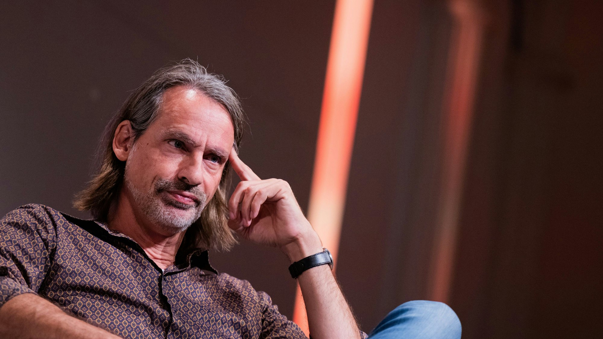 Richard David Precht sitzt auf einem Stuhl und stützt die Hand auf seinem Bein auf.