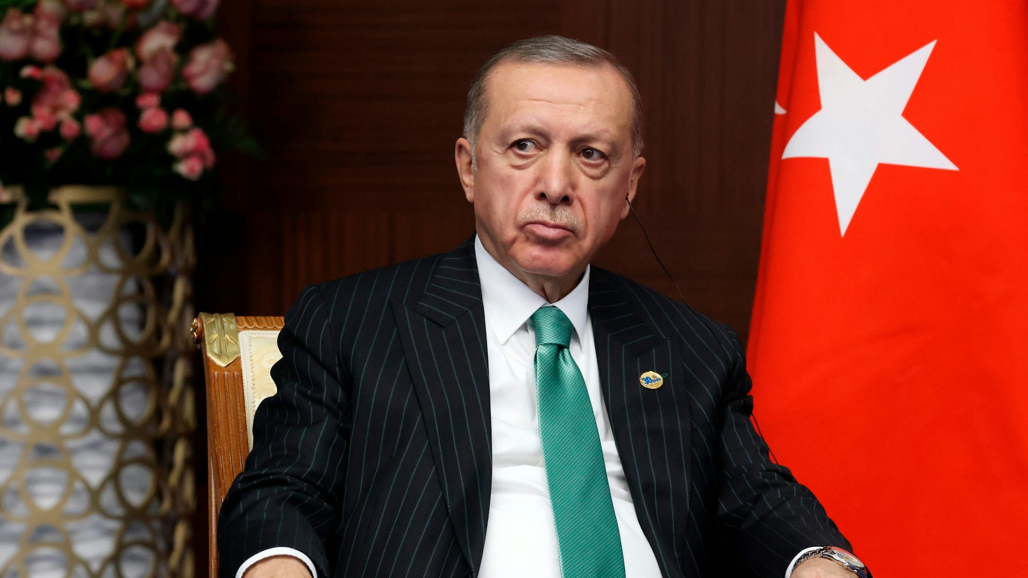 Der türkische Präsident Recep Tayyip Erdogan sitzt auf einem Stuhl und hat einen Kopfhörer im Ohr. Hinter ihm ist die Fahne der türkischen Republik.