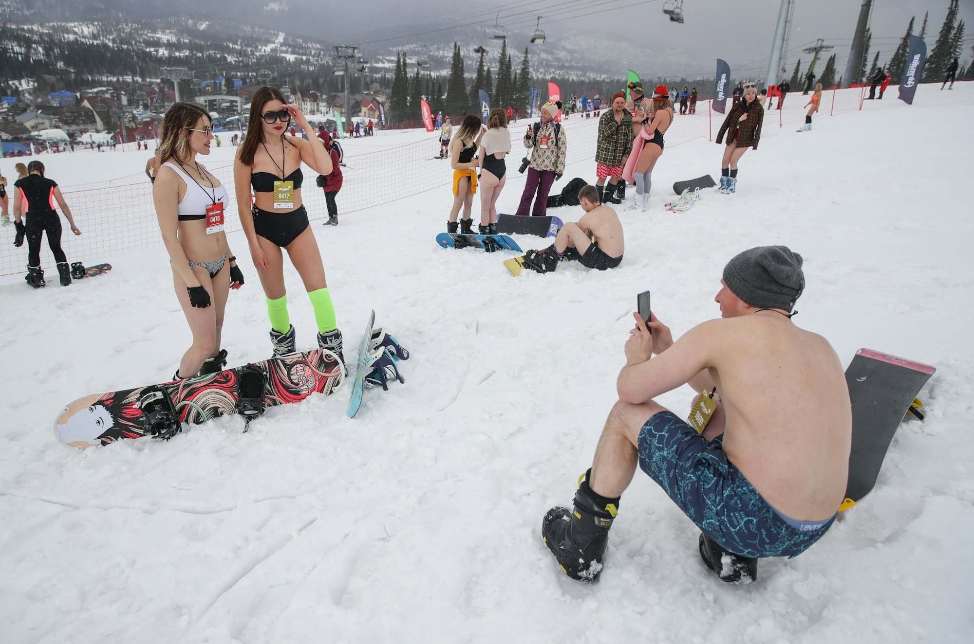 Ein Snowboarder in Badehose fotografiert zwei Frauen, die im Bikini auf einer schneebedeckten Skipiste stehen.