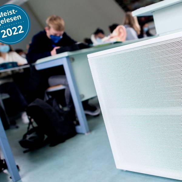 Im Vordergrund sind Luftfilter in einem Klassenraum zu sehen, im Hintergrund Schülerinnen und Schüler mit medizinischen Masken.