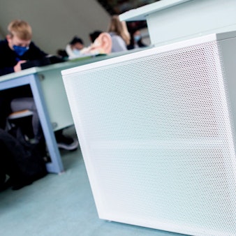 Im Vordergrund sind Luftfilter in einem Klassenraum zu sehen, im Hintergrund Schülerinnen und Schüler mit medizinischen Masken.