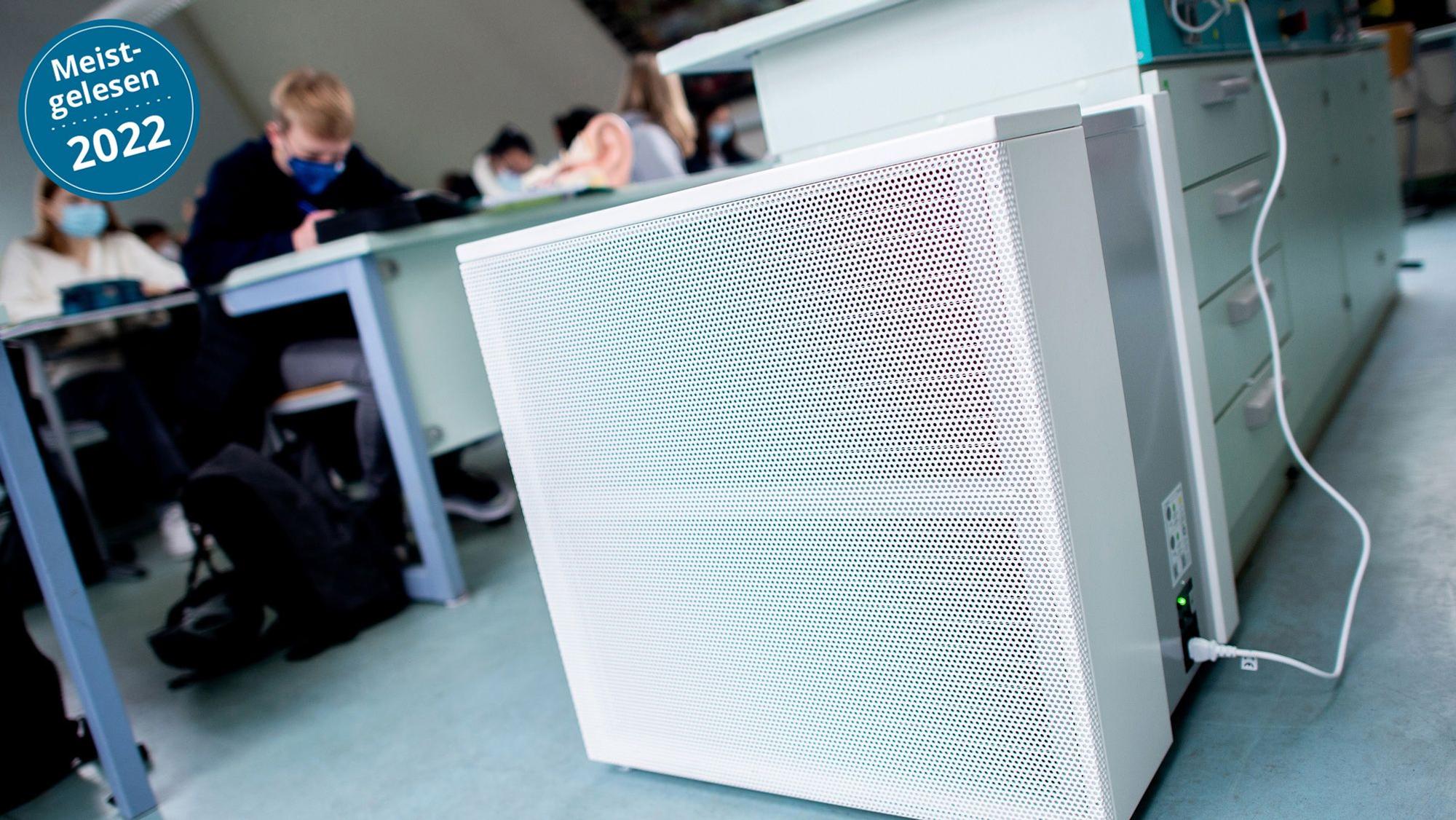Im Vordergrund sind Luftfilter in einem Klassenraum zu sehen, im Hintergrund Schülerinnen und Schüler mit medizinischen Masken.