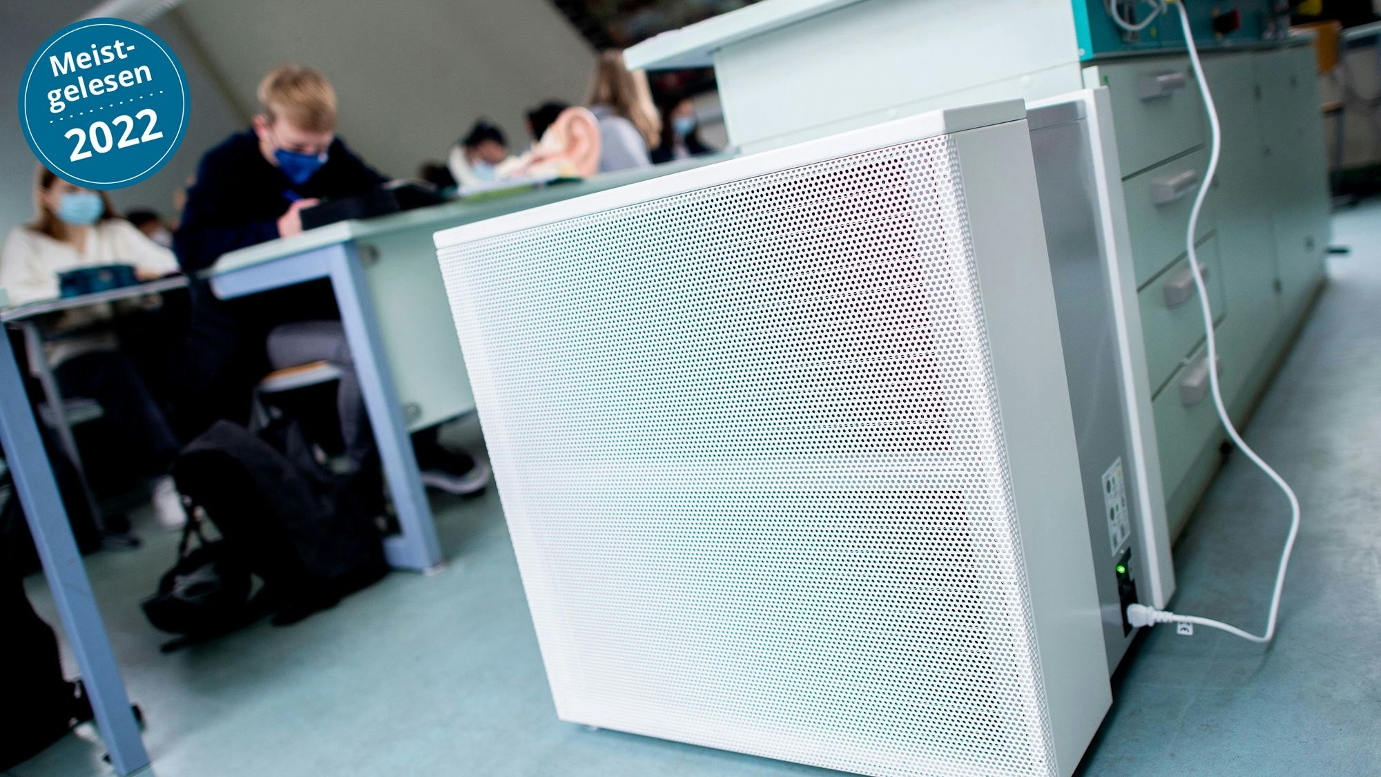 Im Vordergrund sind Luftfilter in einem Klassenraum zu sehen, im Hintergrund Schülerinnen und Schüler mit medizinischen Masken.