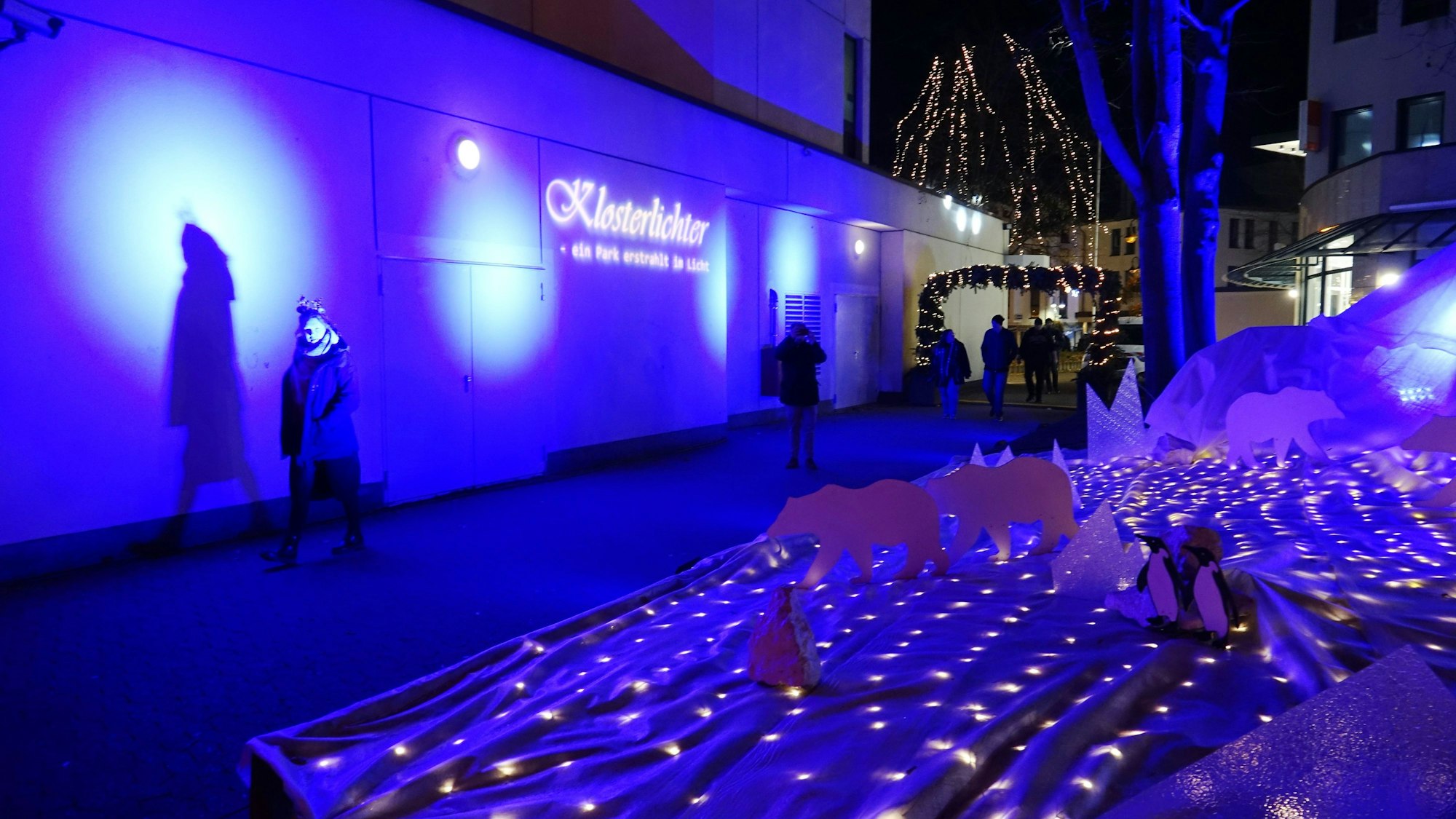 Passanten streben an einer blau angeleuchteten Polarlandschaft in Richtung Klosterlichter.