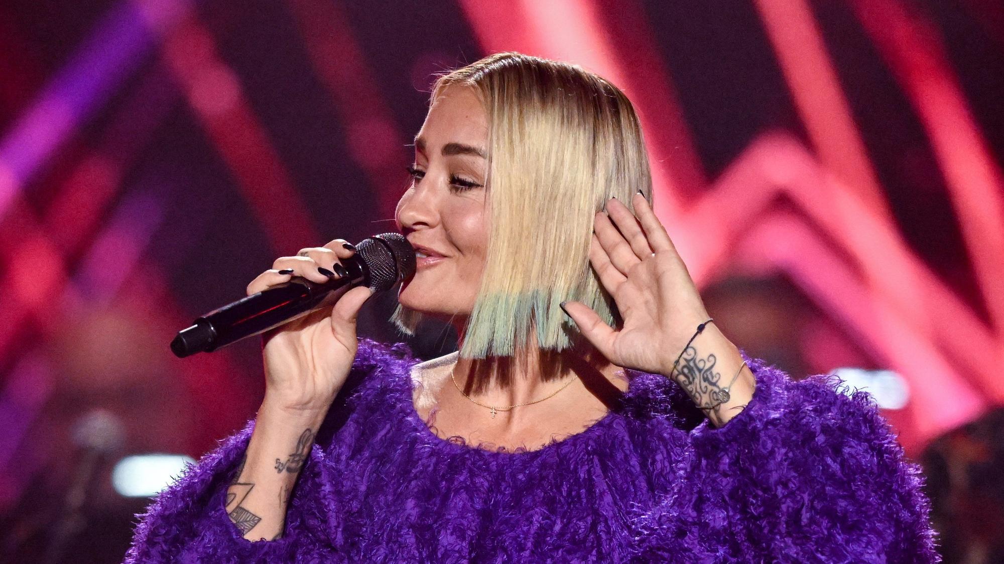 Die deutsche Pop- und Soul-Sängerin Sarah Connor tritt in der ARD-Unterhaltungsshow „Your Songs“ auf. Sie hält lächelnd ihr Mikro an den Mund und ihre andere Hand ans Ohr.