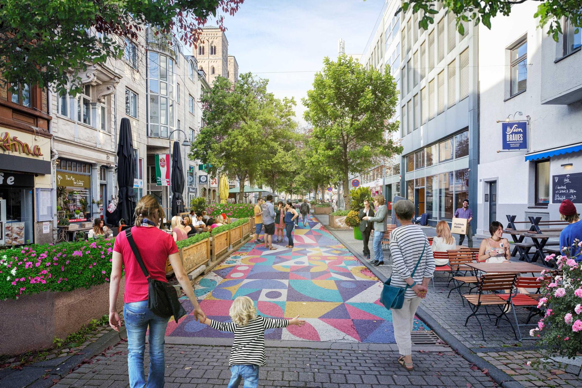 Der große Asphalt auf der Deutzer Freiheit ist bunt angemalt. Fußgänger schlendern über die Straße. Außengastronomie auf ehemaligen Parkplätzen am Straßenrand.