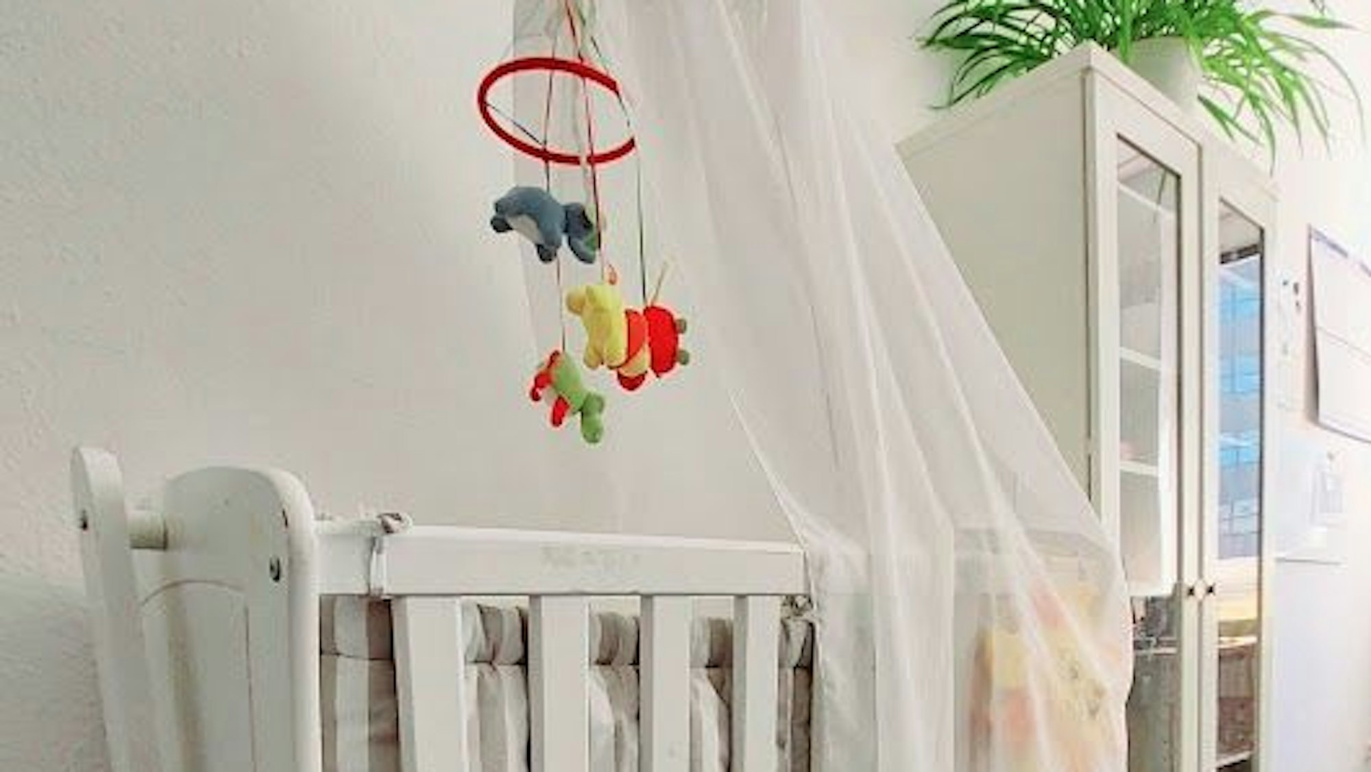 Ein weißes Kindergitterbett mit einem Tier-Mobilé steht neben einem weißen Kleiderschrank.