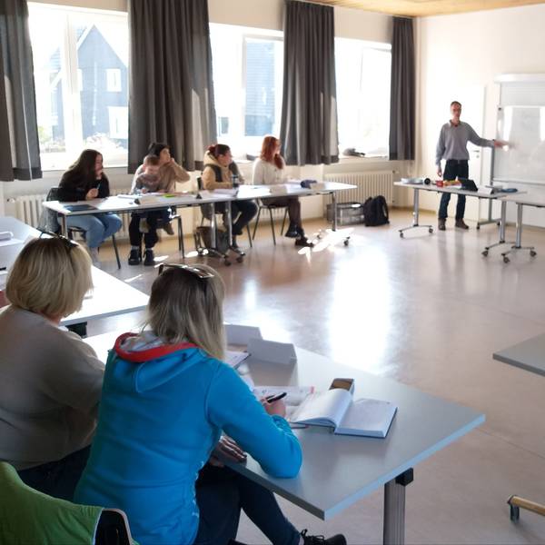Frauen sitzen in einem Raum an Tischen und hören dem Sprachlehrer zu, der vorn an einer weißen Tafel etwas erklärt.