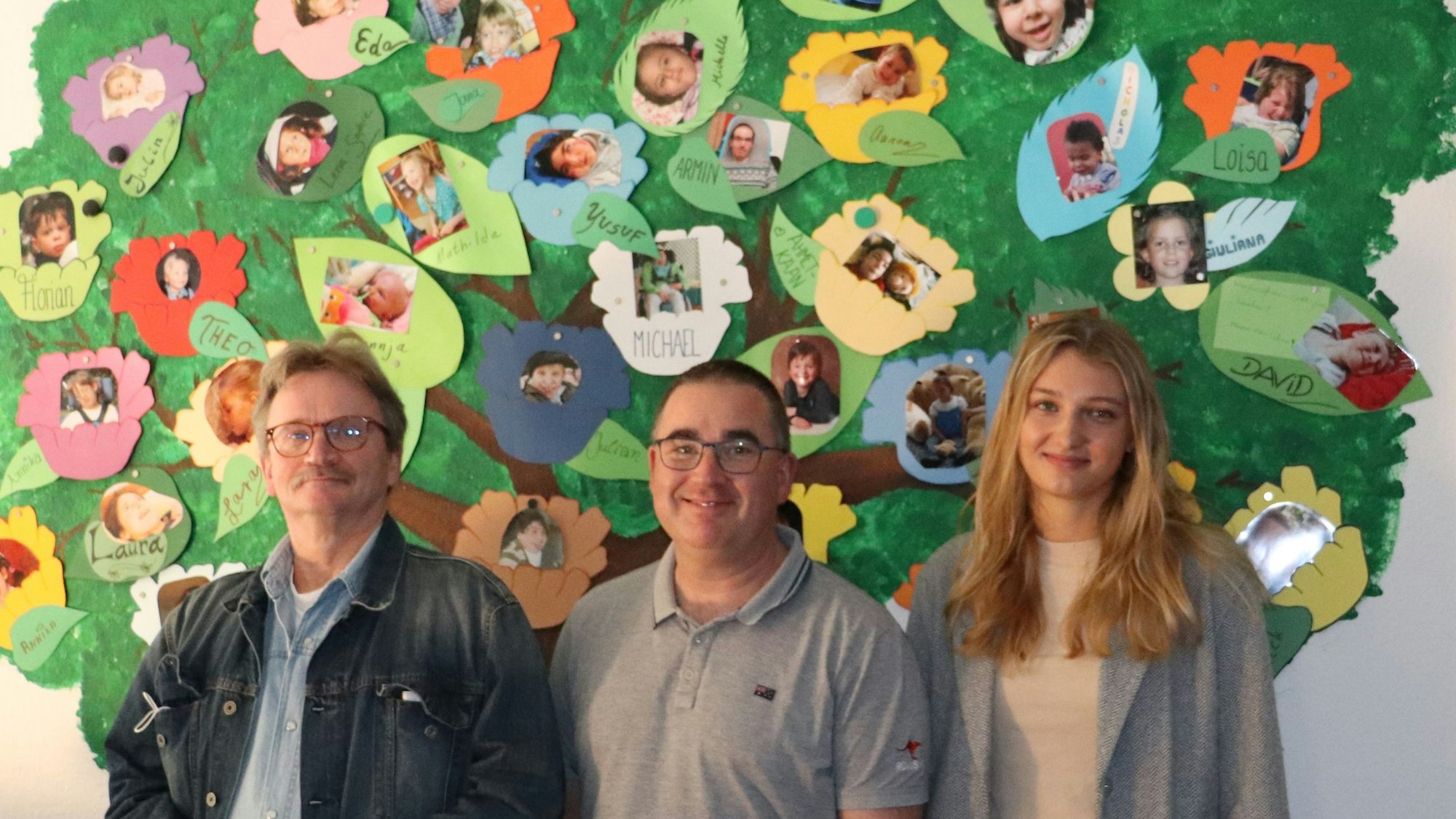 Zwei Mitarbeiter und eine Mitarbeiterin des Ambulanten Kinderhospizdienst Köln stehen vor einer Wand, auf die ein Baum gemalt ist, an dem Fotos von den betreuten Kindern hängen.