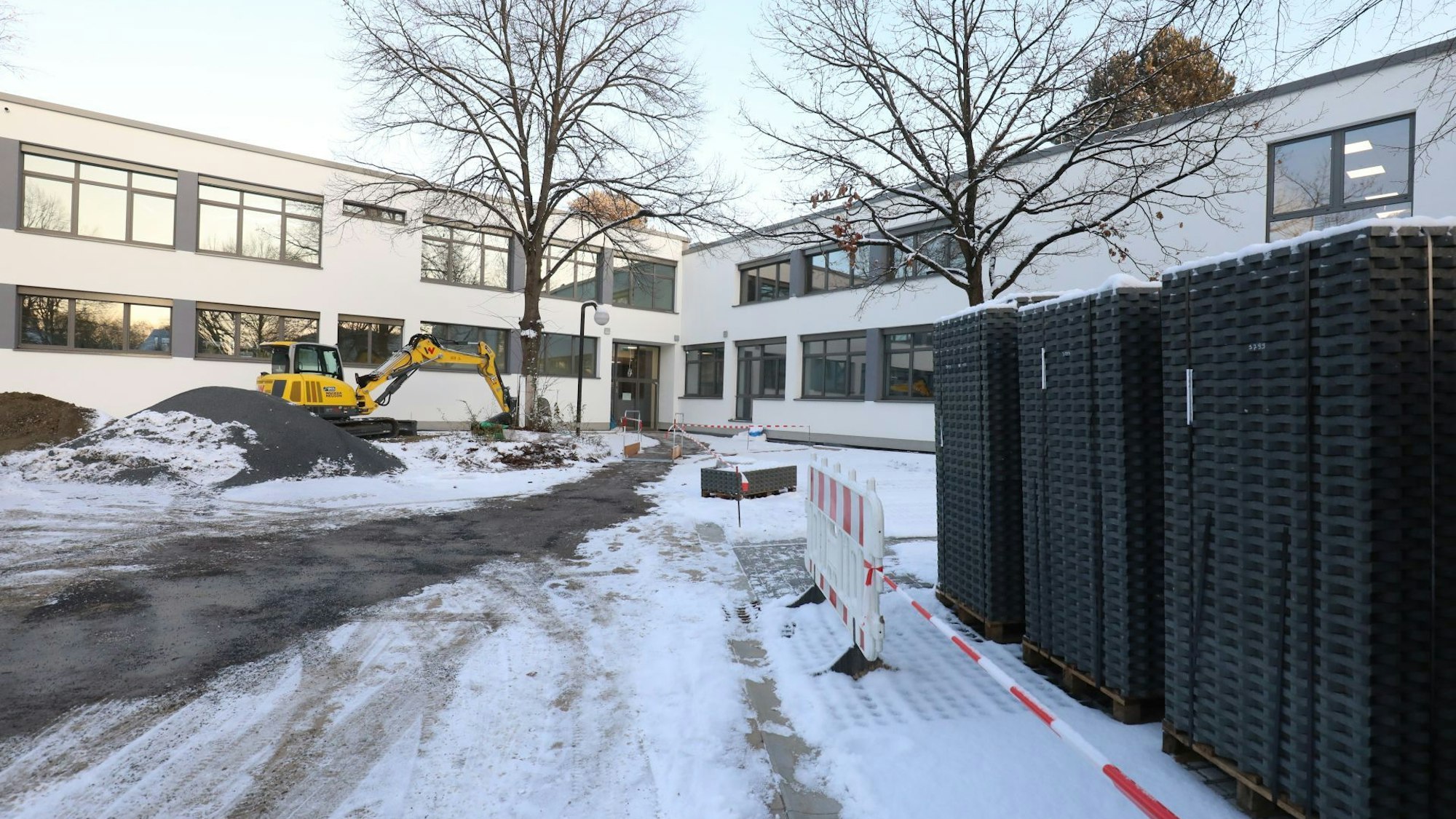 Auf dem mit Schnee bedeckten Vorplatz des L-förmigen Gebäudes stehen ein gelber Bagger und auf Holzpaletten gelagerte schwarze Steine.