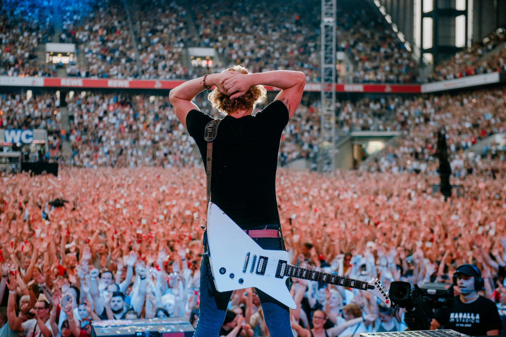 Beim Kasalla-Konzert im Stadion steht Gitarrist Flo Peil mit den Händen überm Kopf vor der jubelnden Menge