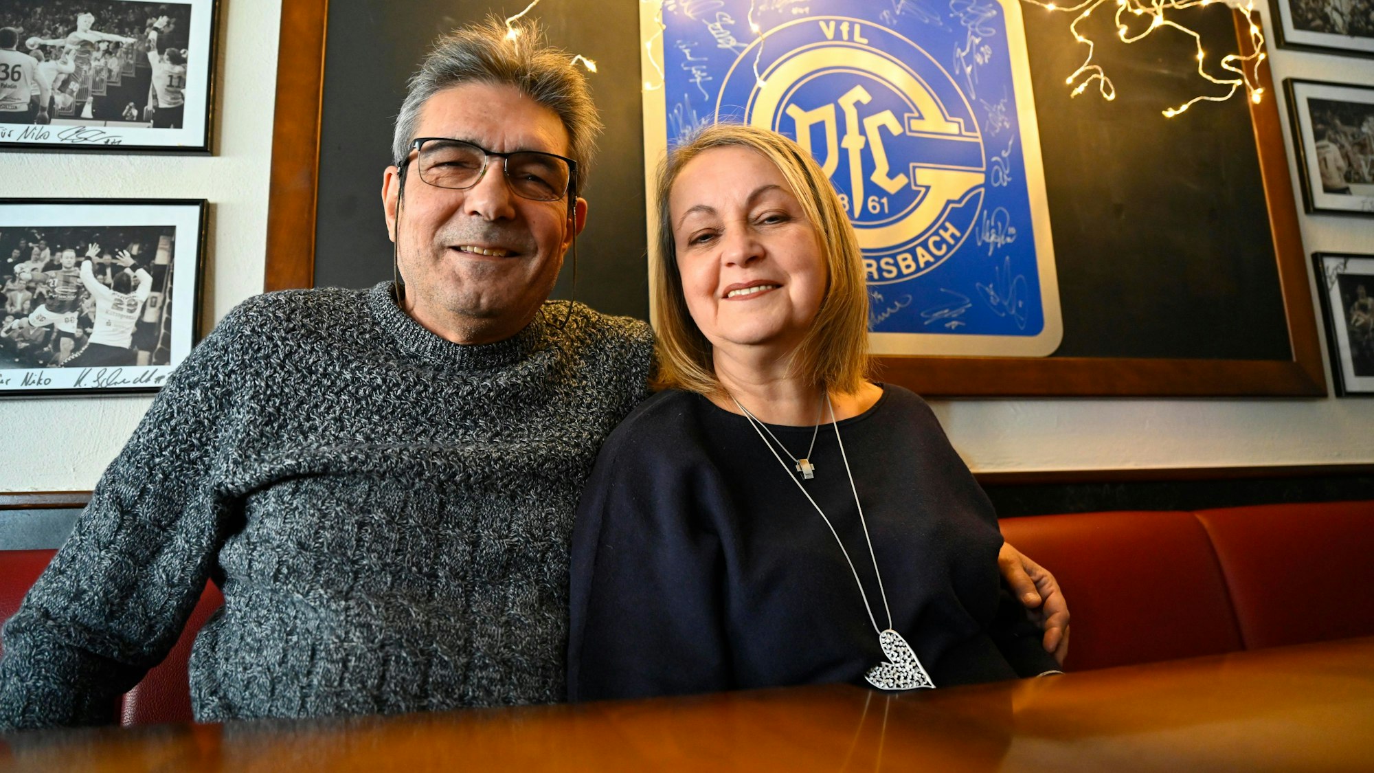 Niko und Christa Argiriou sitzen in ihrer Gastwirtschaft vor dem Logo des Handballvereins VfL Gummersbach.