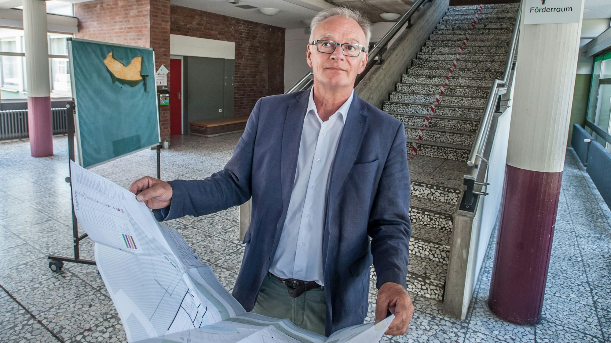 Wolfgang Kalski steht mit Bauplänen in der Hand im Foyer der Grundschule Büscherhof