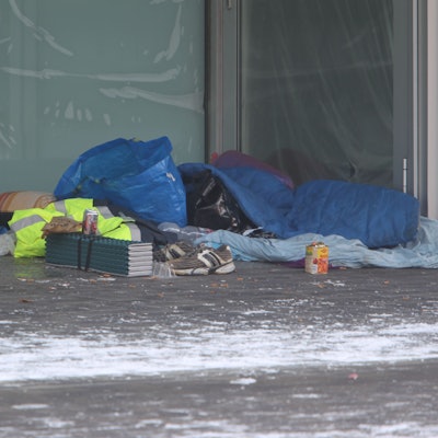 In einer Nische eines Ladenlokals ist ein Matratzenlager mit Plastiktüten , Schlafsack und einem Kissen eingerichtet. Turnschuhe, eine Getränkedose und eine Dose Ravioli stehen davor.