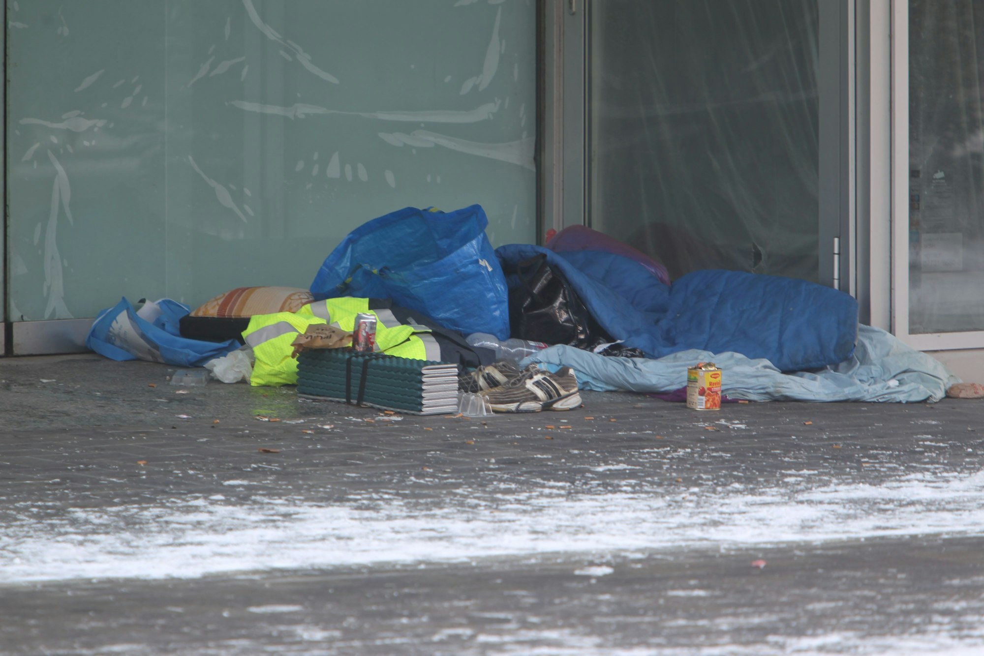 In einer Nische eines Ladenlokals ist ein Matratzenlager mit Plastiktüten , Schlafsack und einem Kissen eingerichtet. Turnschuhe, eine Getränkedose und eine Dose Ravioli stehen davor.
