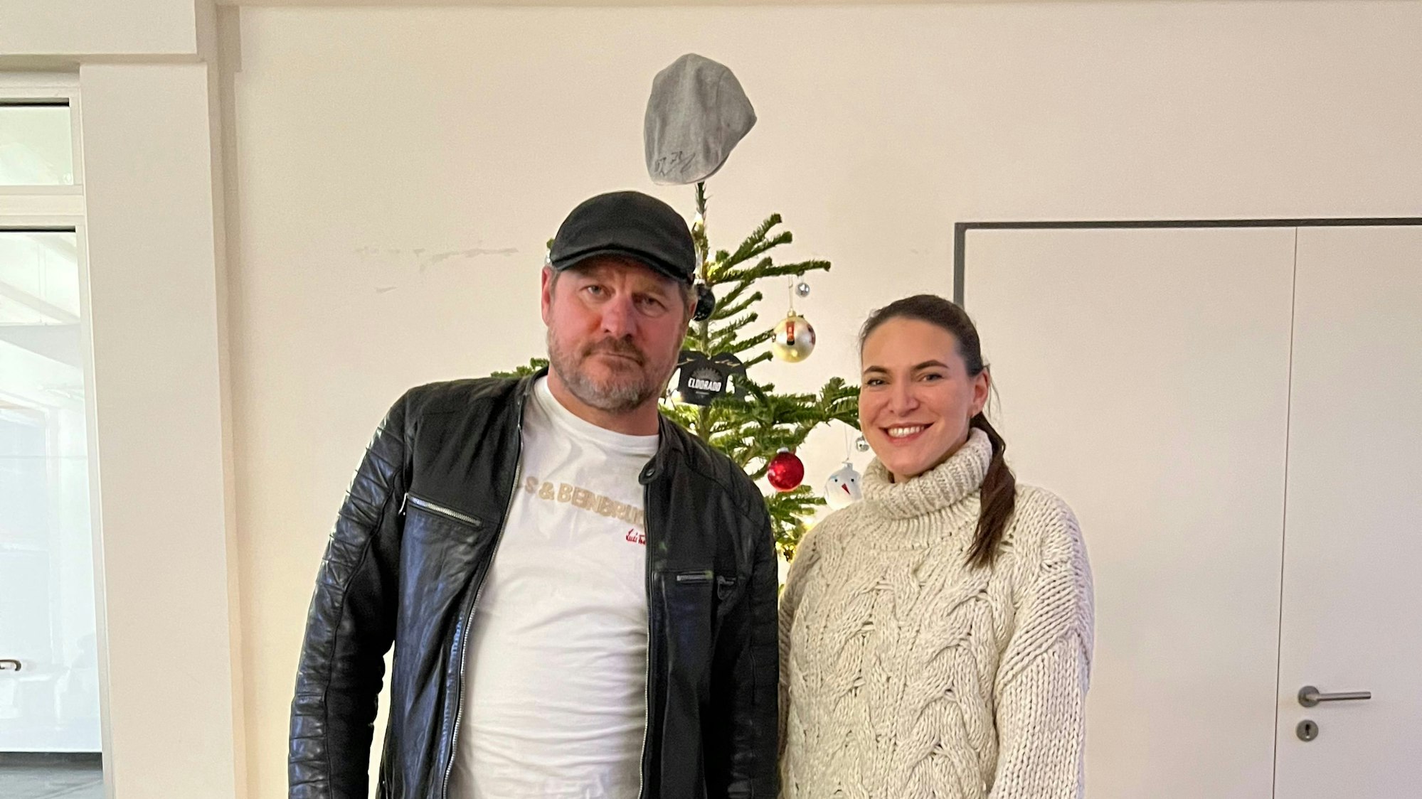 Steffen Baumgart, Trainer des 1. FC Köln, steht mit Lederjacke und Schiebermütze neben Radio-Köln-Moderatorin Pia Pietsch, im weißen Strickpullover. Im Hintergrund ist ein Weihnachtsbaum zu sehen, auf dessen Spitze eine weitere Schiebermütze hängt.