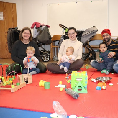 Drei Eltern sitzen mit ihren Babys und einer Diplom-Heilpädagogin auf knallroten Matten, vor ihnen liegt allerhand Spielzeug.