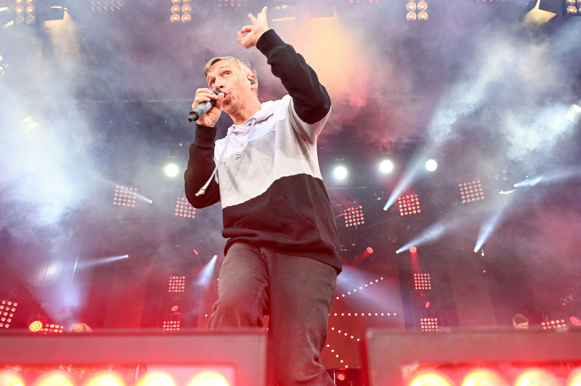 Kasalla-Sänger Bastian Campmann singend auf der Bühne im Gegenlicht von Scheinwerfern und umgeben von Nebel.