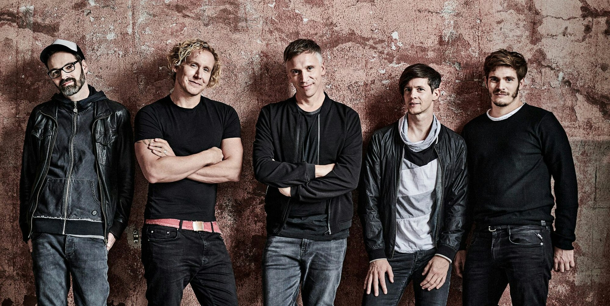 Kasalla in Schwarz und grau gekleidet steht vor eoner schäbigen rötlichen Betonwand. Die Musiker sind Ena Schwiers, Flo Peil, Bastian Campmann, Sebi Wagner und Nils Plum (v.l.)