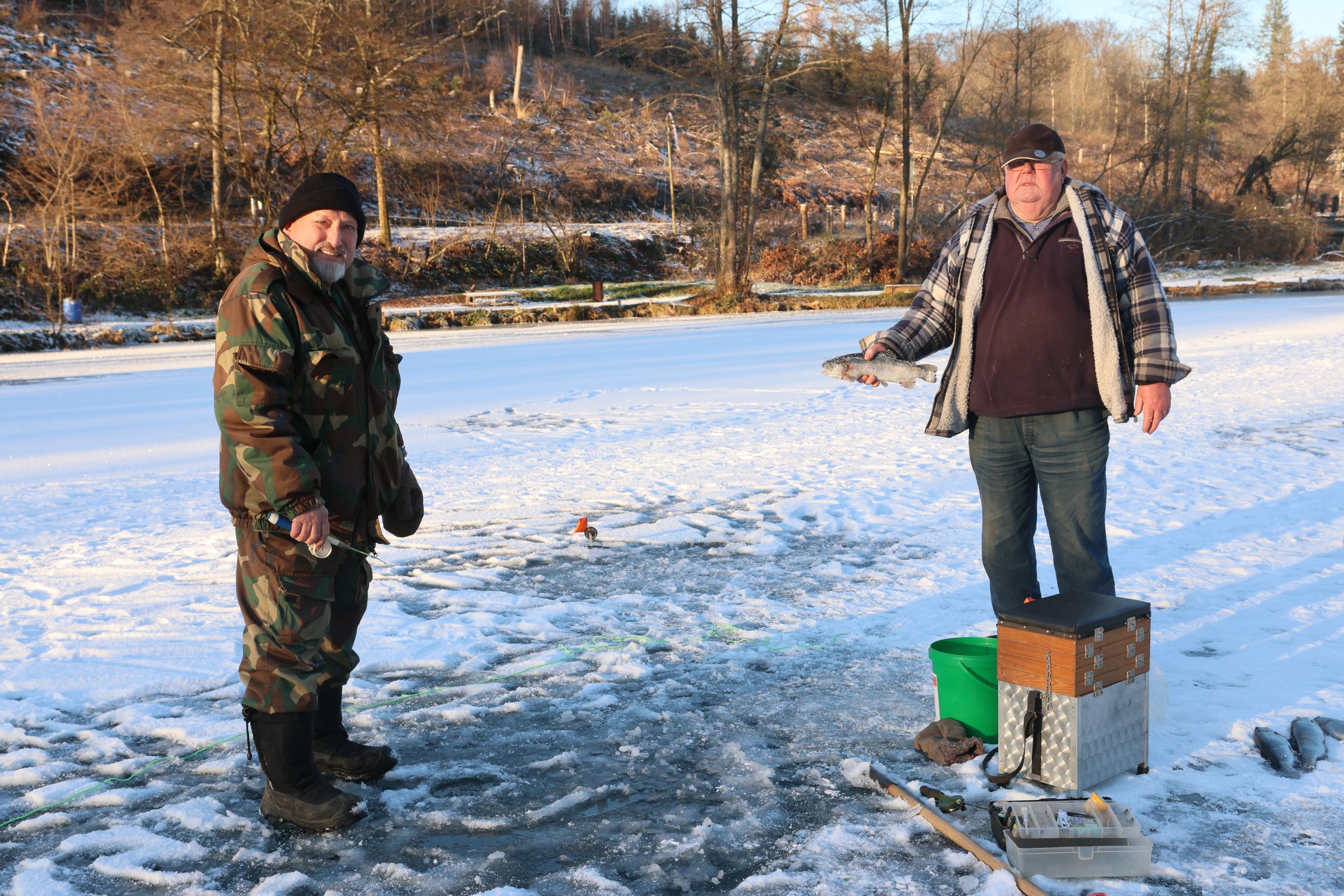 Nikolai und Eduard Peters stehen nebeneinander auf einem zugefrorenen See. Neben ihnen liegen Utensilien, die sie zum Eisangeln benötigen.