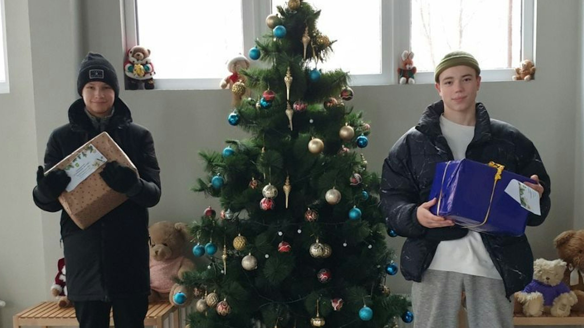 Zwei Kinder stehen mit Geschenken neben einem geschmückten Baum.