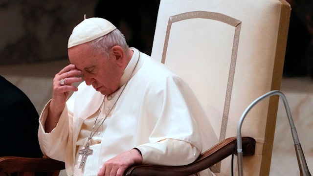 Papst Franziskus sitzt in einem Stuhl und fasst sich an den Kopf. Um seinen Hals hängt ein silbernes Kreuz.