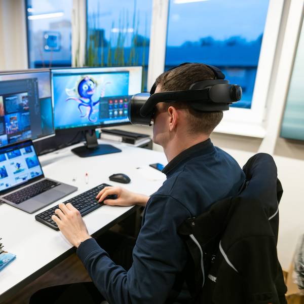 Ein Mann sitzt mit VR-Brille an einem Arbeitsplatz mit mehreren PC-Bildschirmen.