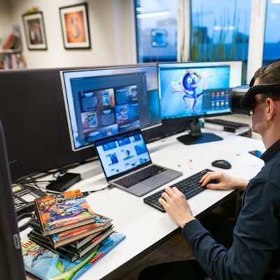 Ein Mann sitzt mit VR-Brille an einem Arbeitsplatz mit mehreren PC-Bildschirmen.