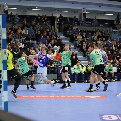 Man sieht eine Spielsituation, der junge Handballer Finn Schroven setzt zum Wurf auf das Tor an. Um ihn herum stehen Spieler in anderen Trikots, im Hintergrund sieht man die Tribüne.