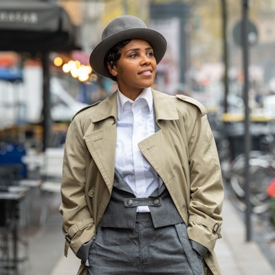 Alice Francis steht in Anzughose, Bluse, Trenchcoat und mit Hut auf der Aachener Straße in Köln.