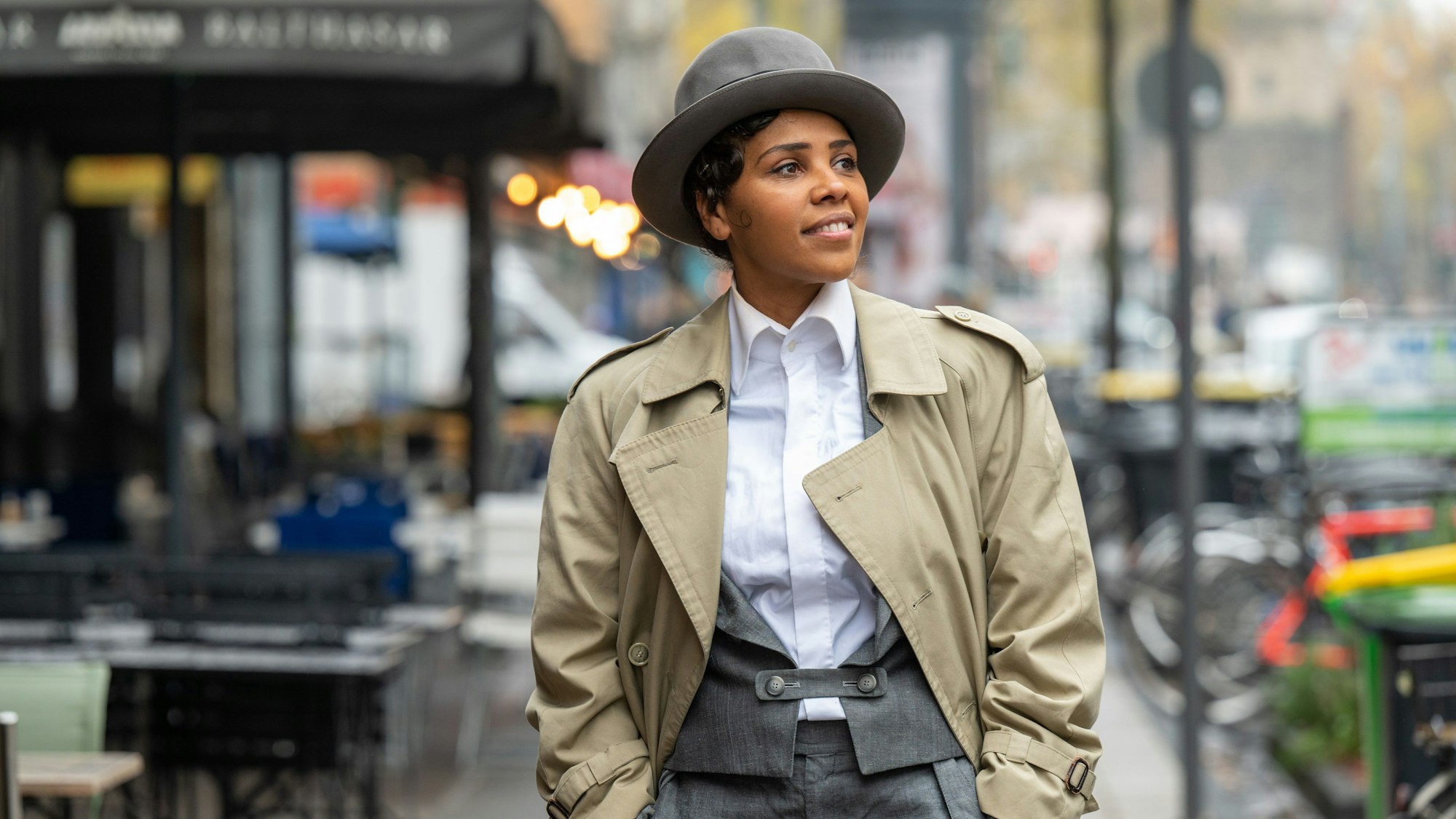 Alice Francis steht in Anzughose, Bluse, Trenchcoat und mit Hut auf der Aachener Straße in Köln.