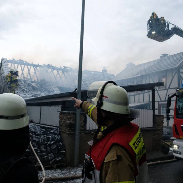 Zwei Einsatzkräfte sowie ein Drehleiter-Fahrzug der Feuerwehr stehen vor dem gelöschten Gebäude.
