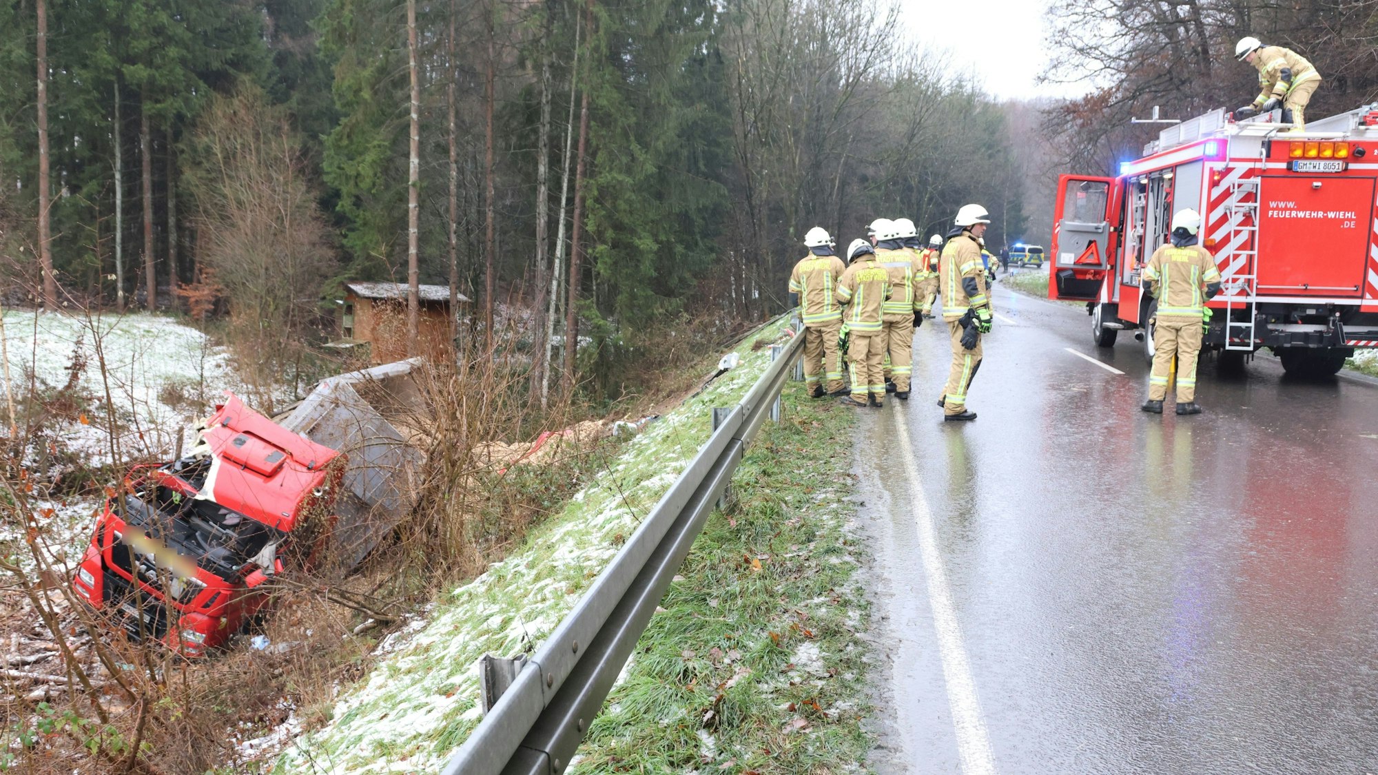 Feuerwehrleute stehen auf der Straße, links hinter der Leitplanke liegt der verunglückte LKW. Die Polizei sperrt die Straße ab.