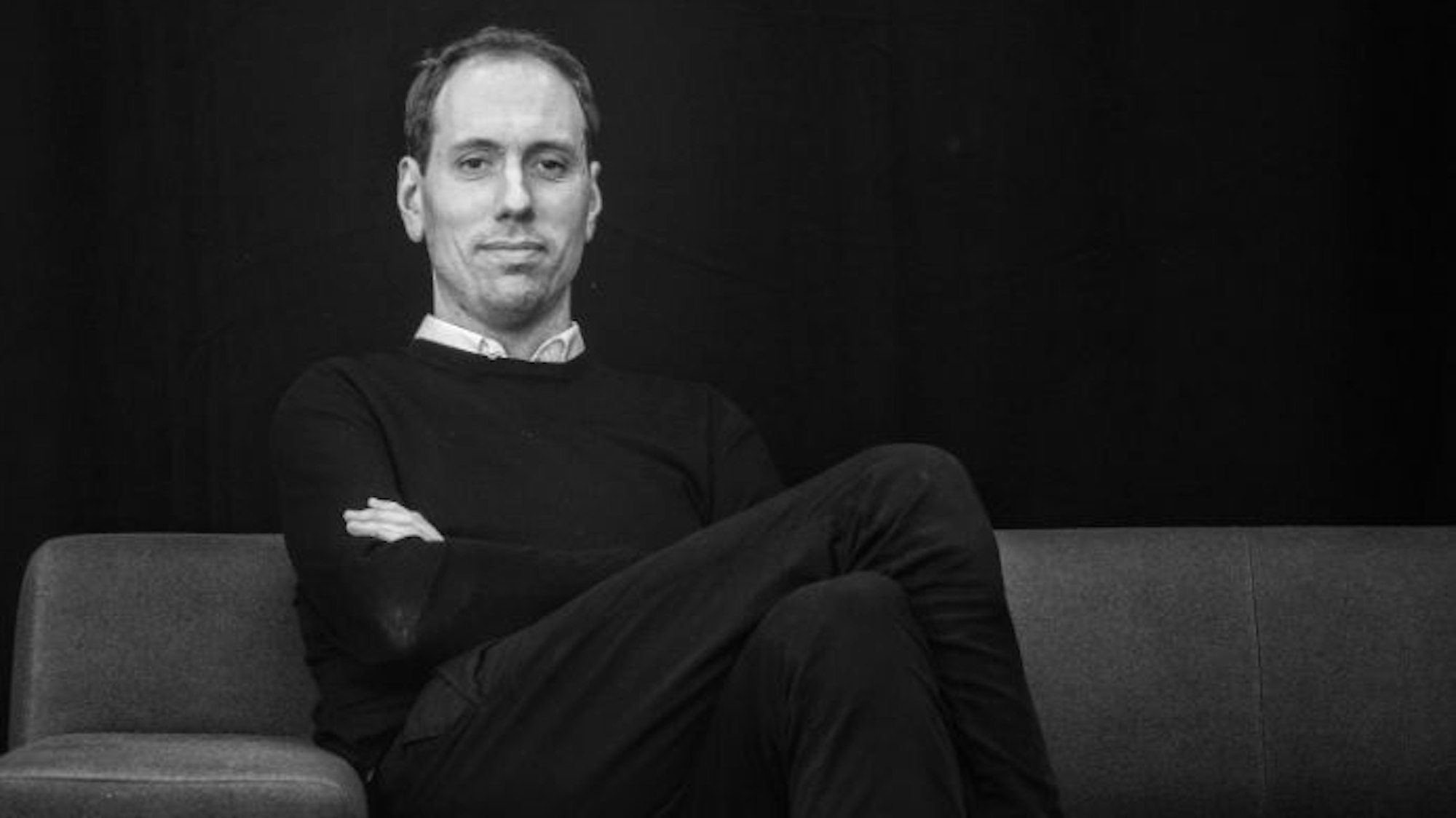 Der Lionel Meunier auf einer Couch, die Beine übereinandergeschlagen und die Arme überkreuzt. Das Foto ist schwarz-weiß.