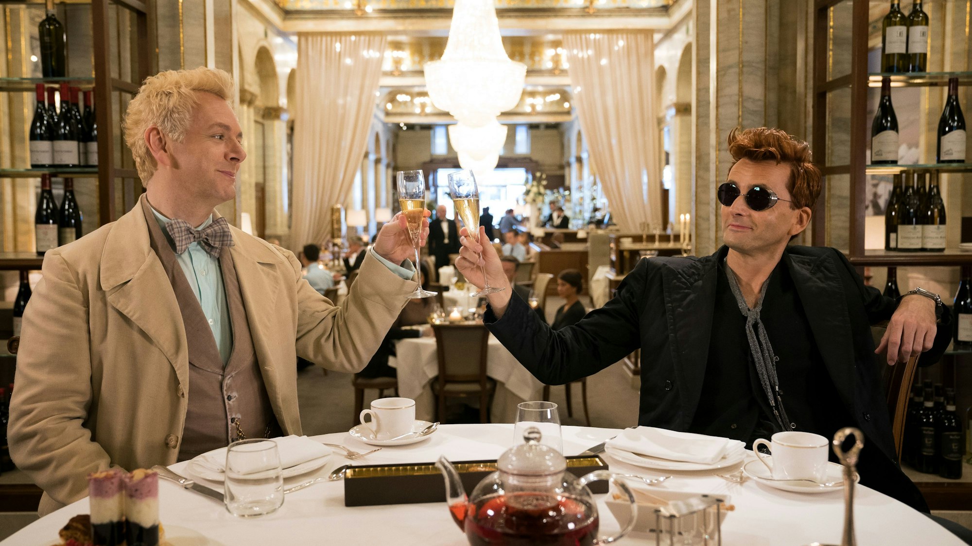 Michael Sheen (l) als Engel Erziraphael und David Tennant als Dämon Crowley in einer Szene der Comedy-Serie „Good Omens“. Sie stoßen mit Sekt an und lächeln.