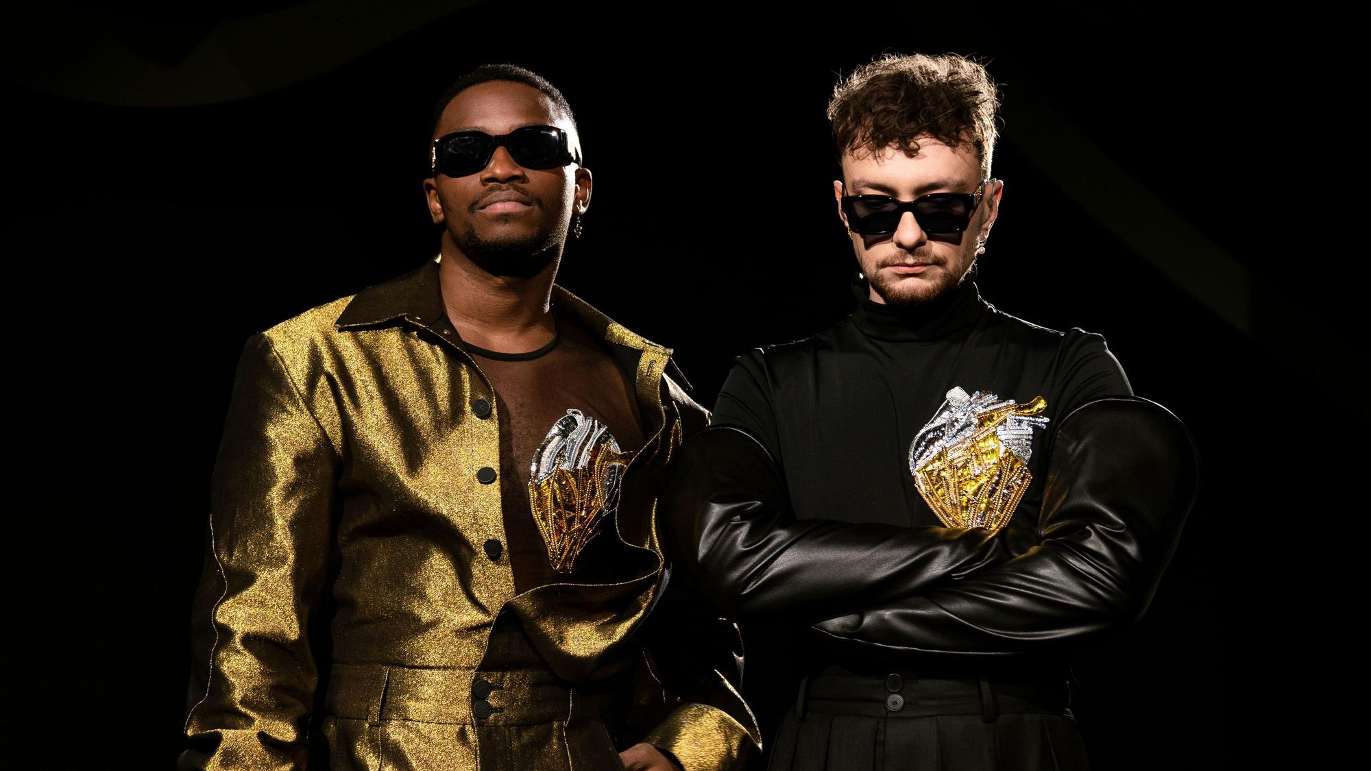 Jeffery Kenny (l) und Andrij Guzuljak von der Band TVORCHI in einer U-Bahn-Station. Sie tragen goldene bzw. schwarze Bühnenoutfits und dunkle Sonnenbrillen.