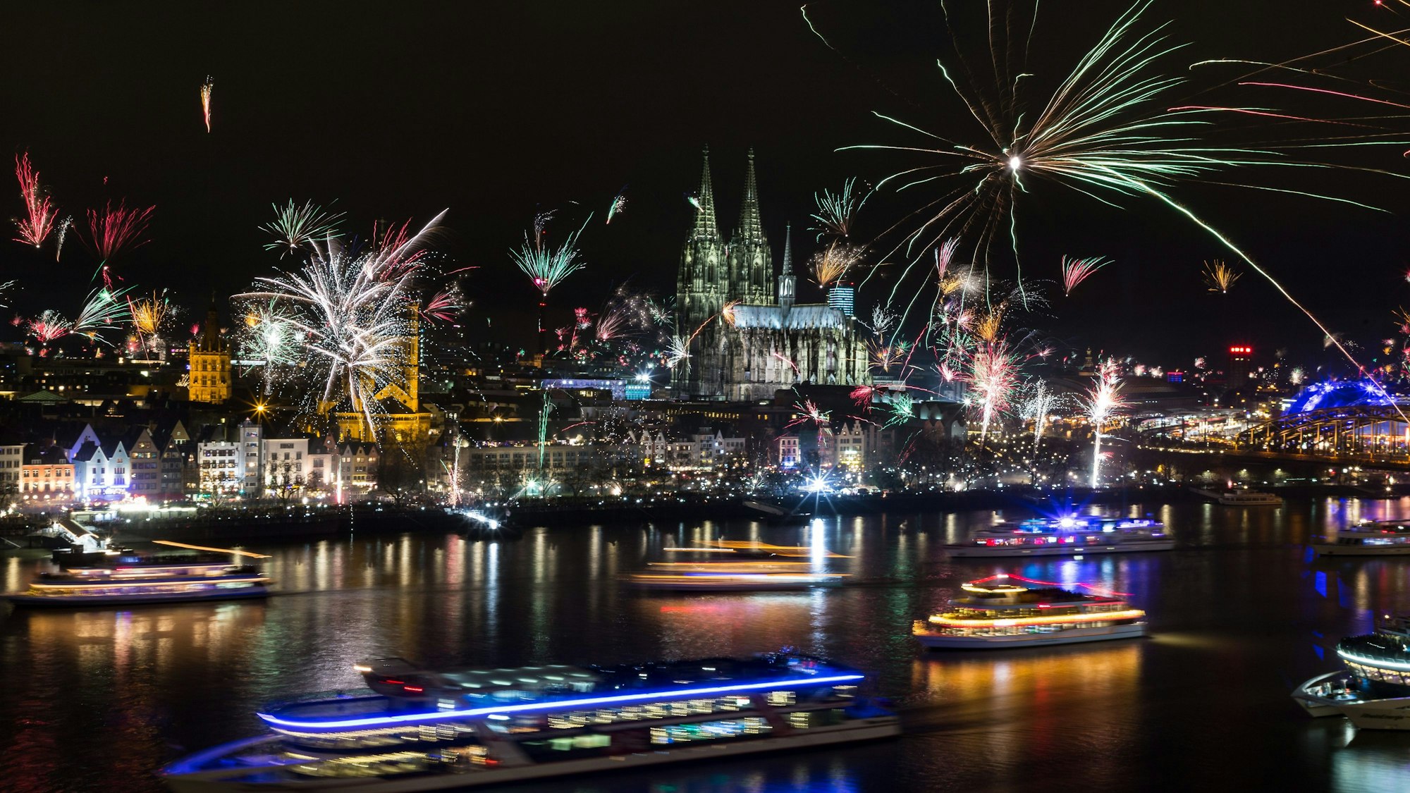 Zahlreiche Feuerwerksraketen explodieren rund um den Kölner Dom. Auf dem Rhein sind erleuchtete Schiffe unterwegs.