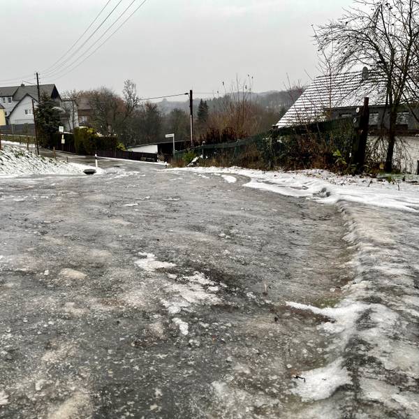 Eine zentimeterdicke Eisschicht liegt auf einer abschüssigen Straße im Hennefer Ortsteil Hanf. Im Hintergrund sind Häuser zu sehen.