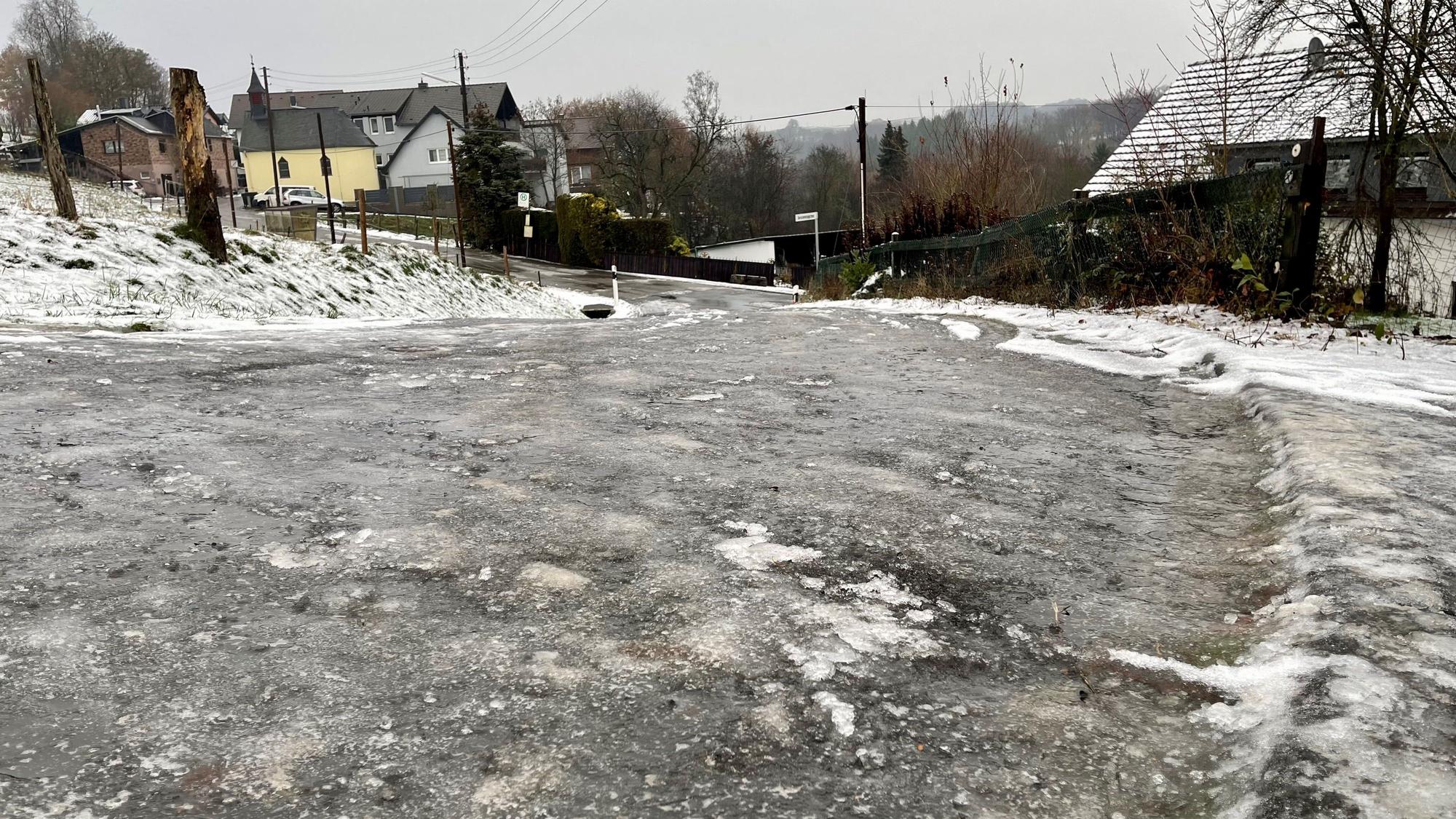 Eine zentimeterdicke Eisschicht liegt auf einer abschüssigen Straße im Hennefer Ortsteil Hanf. Im Hintergrund sind Häuser zu sehen.