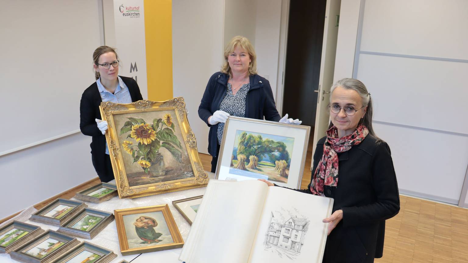 Zeichnungen und Gemälde des Euskirchener Malers Willy Loos, die Enkelin Iris Pütz an Leiterin Heike Lützenkirchen und deren Mitarbeiterin Jennifer Kirchhoff überreicht, sind auf einem Tisch ausgebreitet.