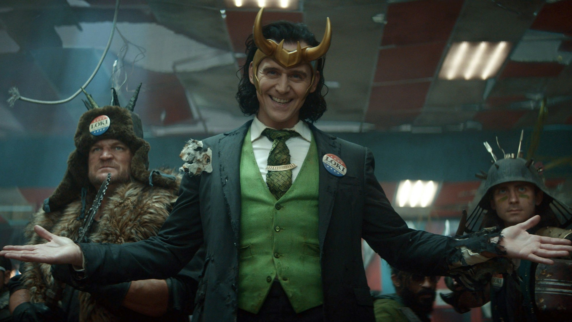 Loki (Tom Hiddleston, M) in einer Szene der Marvel-Serie „Loki“. Er steht vorne und grinst frech. Hinter ihm sind weitere Figuren der Serie.
