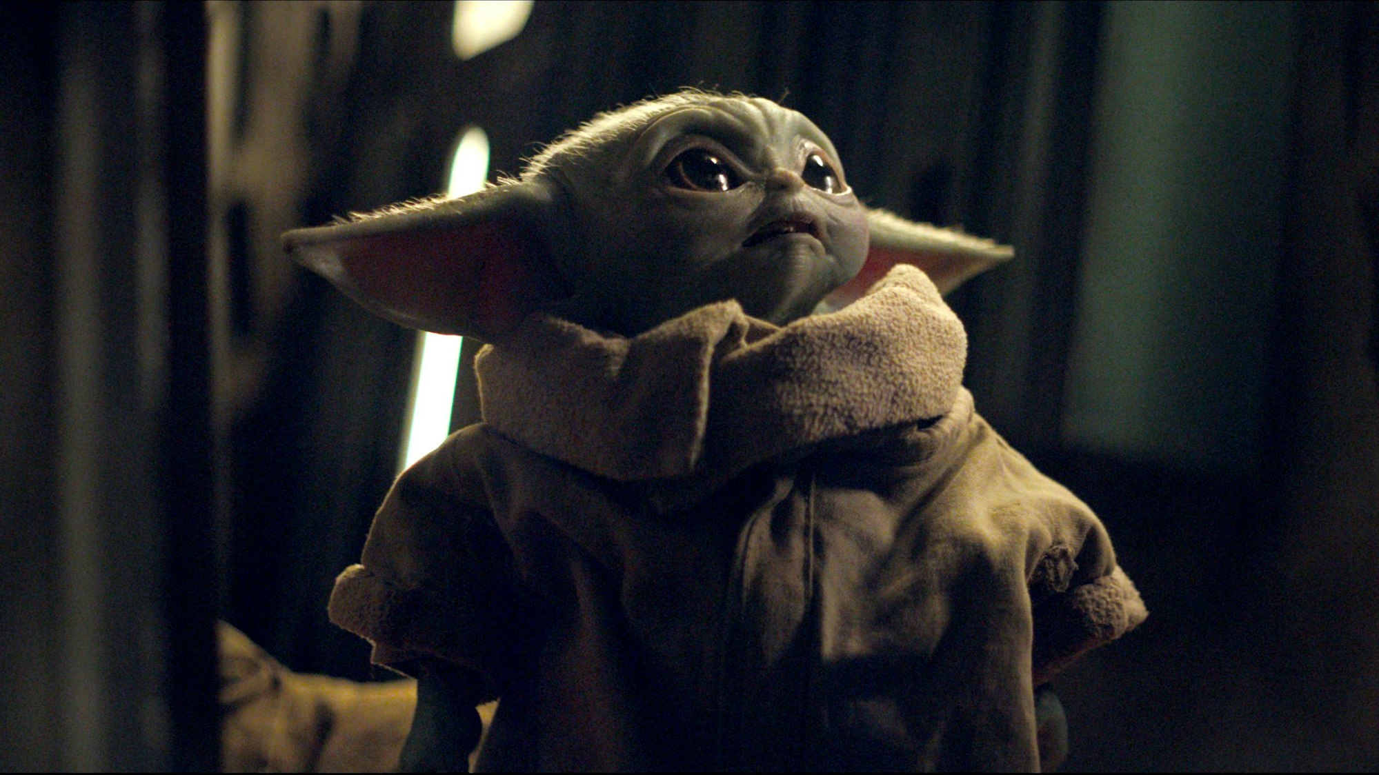 Die Figur ‚Das Kind‘ (Baby Yoda) in einer Szene aus „The Mandalorian“.Das Wesen hat große Kulleraugen, weiche Öhrchen und einen zarten Haar-Flaum auf dem Kopf.