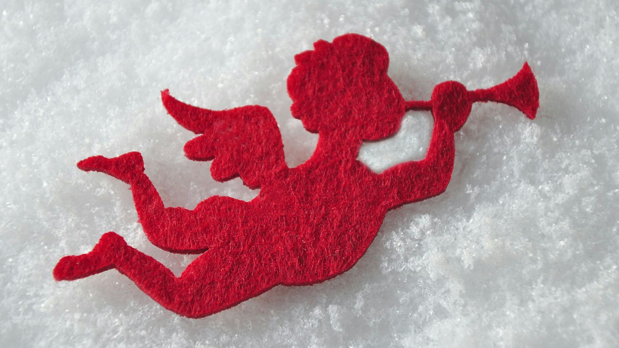 Ein roter Filzengel bläst in eine Trompete, im Hintergrund Schnee.
