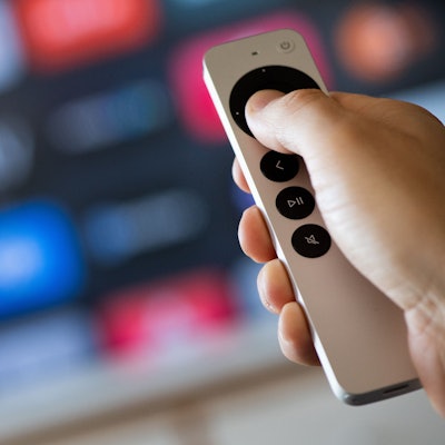 Vor einem verschwommenen Fernsehbildschirm hält eine Hand eine Fernbedienung.