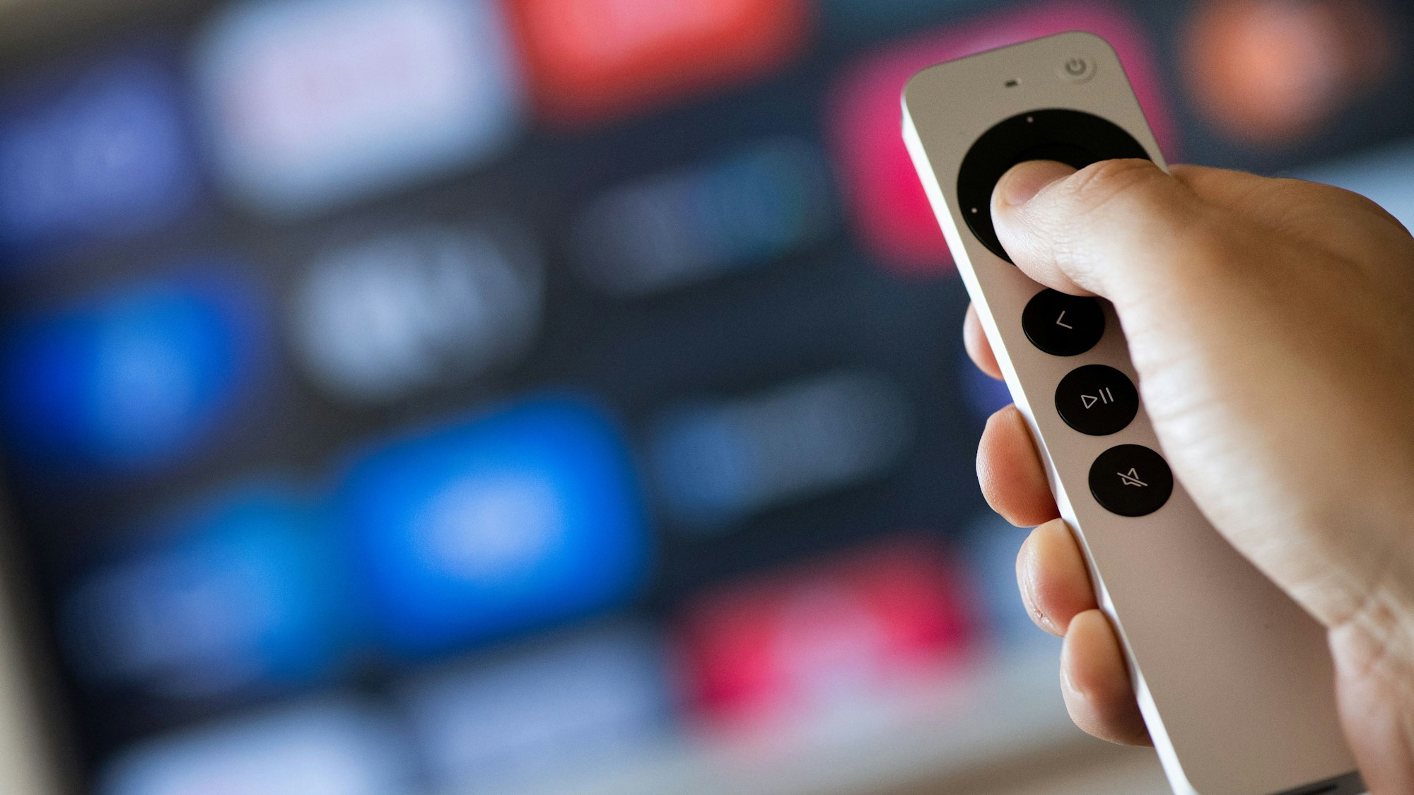 Vor einem verschwommenen Fernsehbildschirm hält eine Hand eine Fernbedienung.