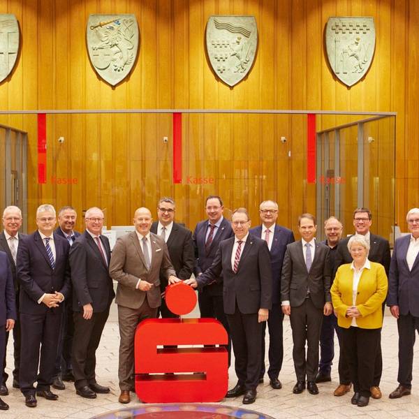 Angestellte der Sparkassen aus Köln und Radevormwald/Hückeswagen stehen in der Hauptstelle der Kreissparkasse Köln vor einem Sparkassen-Logo
