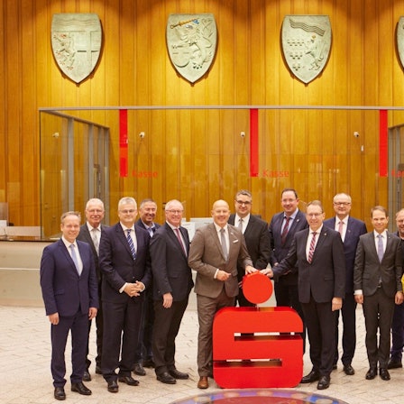 Angestellte der Sparkassen aus Köln und Radevormwald/Hückeswagen stehen in der Hauptstelle der Kreissparkasse Köln vor einem Sparkassen-Logo